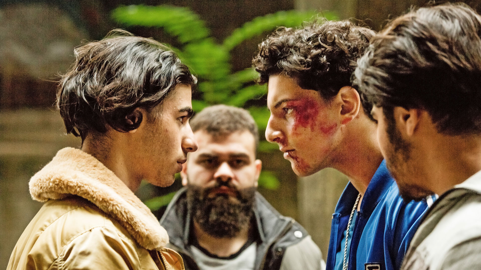 Filmszene: 4 junge Männer schauen sich ernst an. Ein Mann blutet im Gesicht.