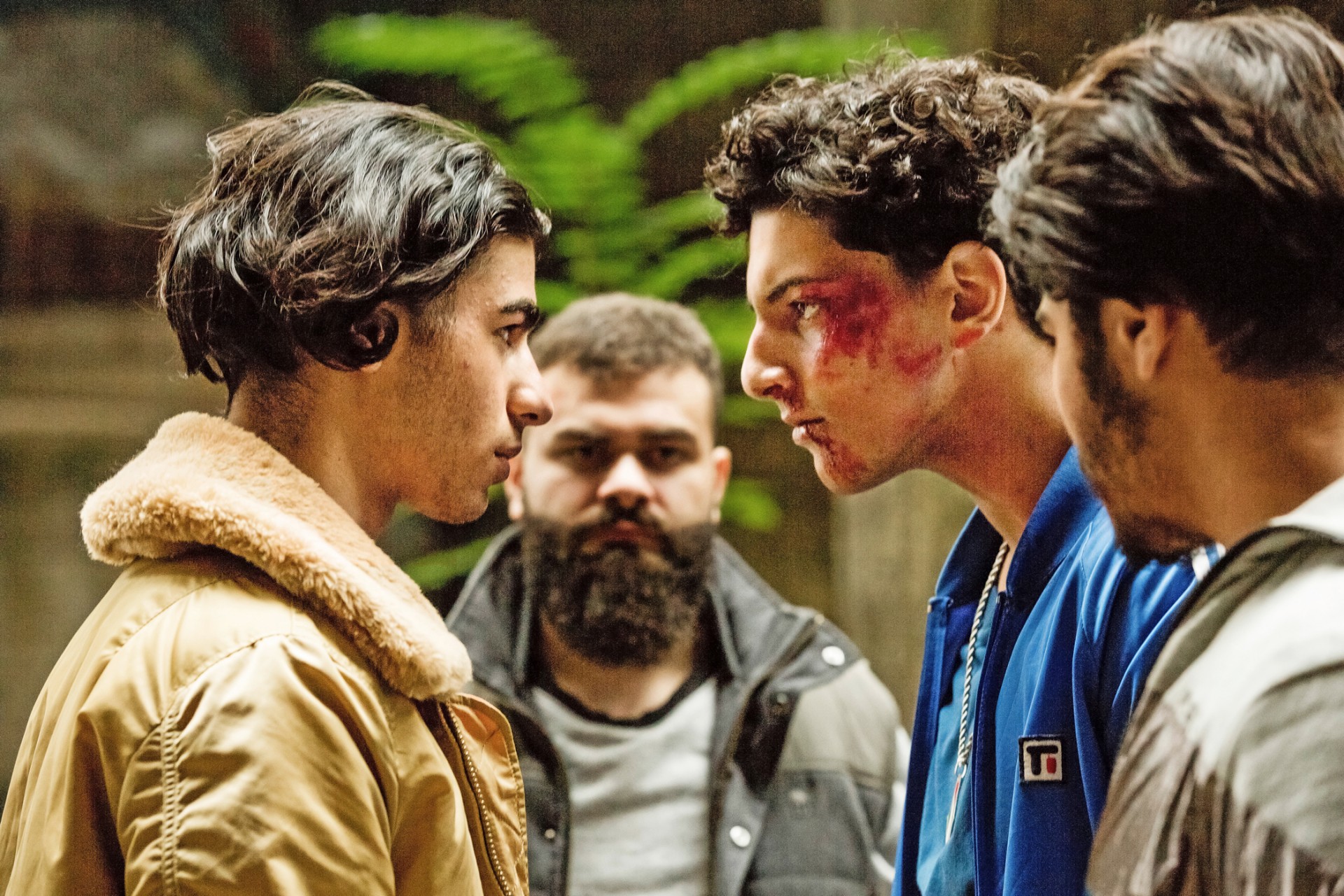 Filmszene: 4 junge Männer schauen sich ernst an. Ein Mann blutet im Gesicht.